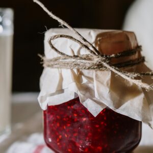 A close-up of homemade raspberry jam in a rustic glass jar on a white table.